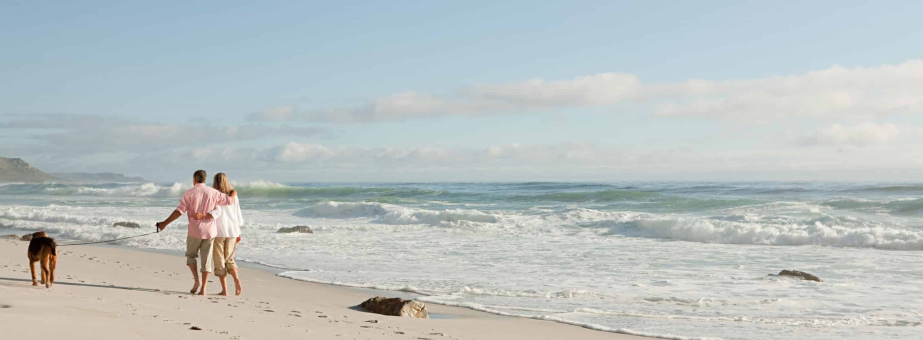 A middle-aged couple walk their dog along a beach.